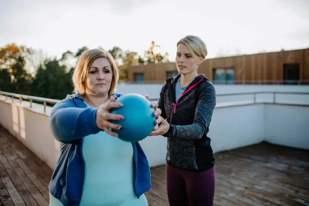Woman exercising with a ball while being helped by a trainer | Featured image for the Blood Pressure Exercises page at Pivotal Motion Physiotherapy.